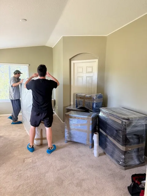 Two Sun City Moving crew members in shoe covers wrapping furniture, with wrapped pieces ready to go